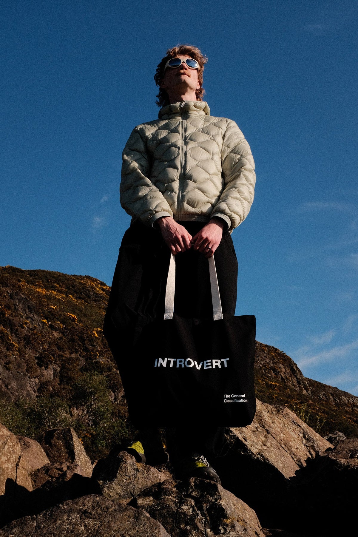 Person standing on a rocky outcrop holding a bag with 'INTROVERT' text against a blue sky.