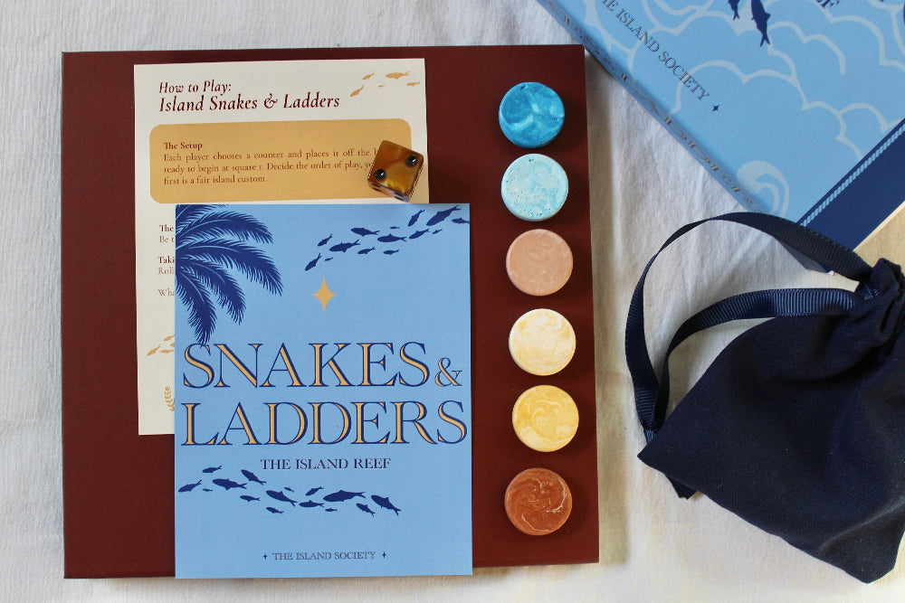 Overhead view of The Island Reef Snakes and Ladders game with blue illustrated board, playing pieces, dice, navy linen bag and box.