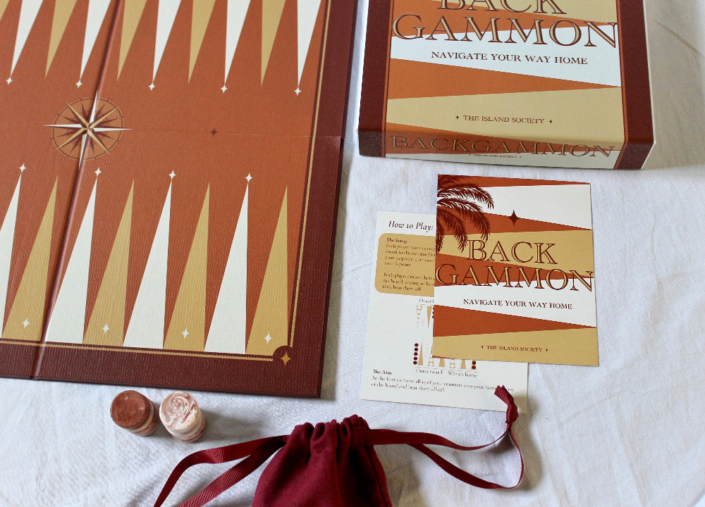 The Island Society backgammon set showing the illustrated board, branded box, game instructions and burgundy drawstring bag.