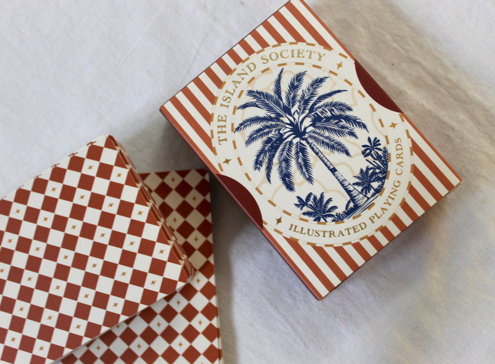 A vintage deck of illustrated playing cards from 'The Island Society' shown with its striped and checkered box on a light fabric background.