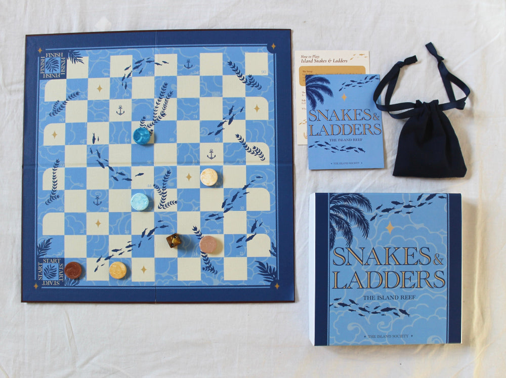 The Island Society Snakes and Ladders game with illustrated blue packaging, handmade Jesmonite counters, dice and navy drawstring bag.