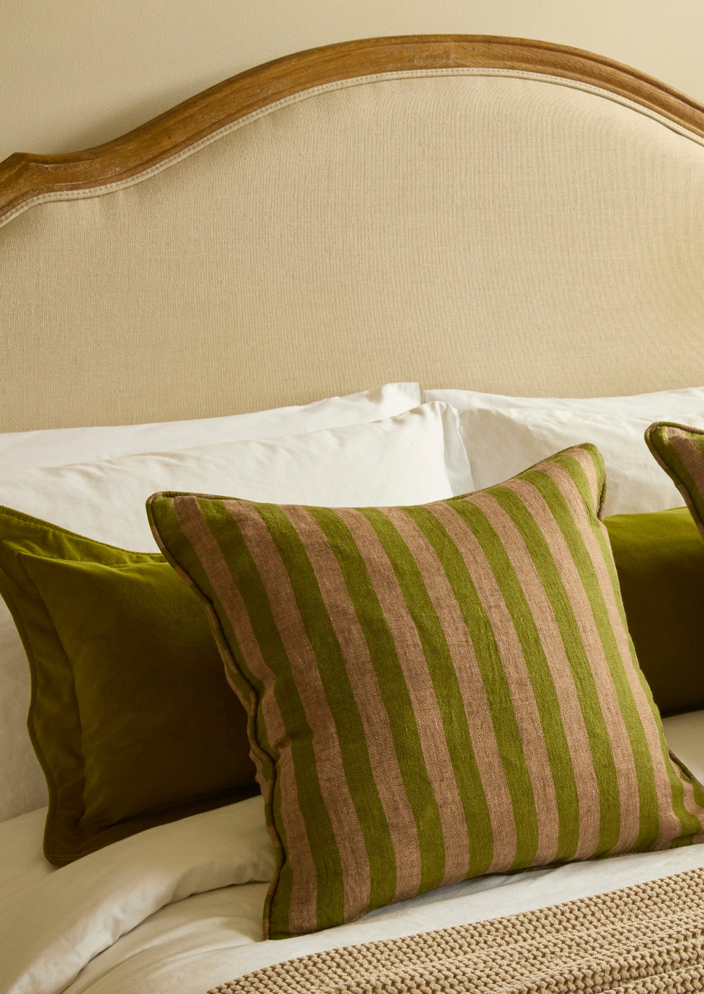 A styled bedroom scene showing a beige upholstered headboard with layered bedding and decorative pillows, including a central striped olive green and tan pillow.