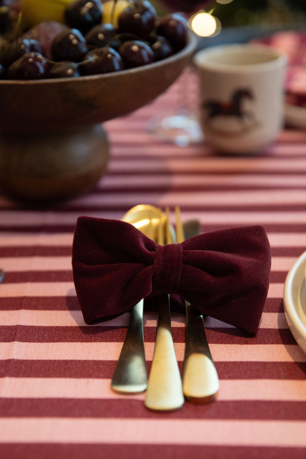 A burgundy velvet bow tie placed around three pieces of silver cutlery on a pink and burgundy striped tablecloth, with a wooden bowl of dark grapes and a blurred mug in the background.