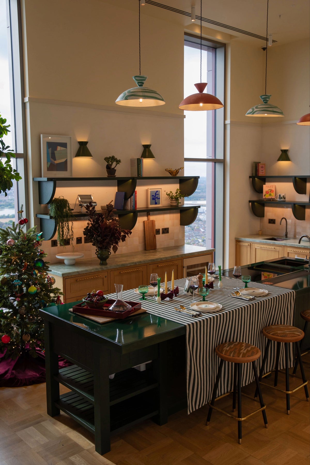 A styled kitchen interior with a dark green island table set with a striped table runner, glassware, and candles, surrounded by wooden stools. The background features a kitchen with wooden cabinets, floating shelves, and colorful pendant lights.