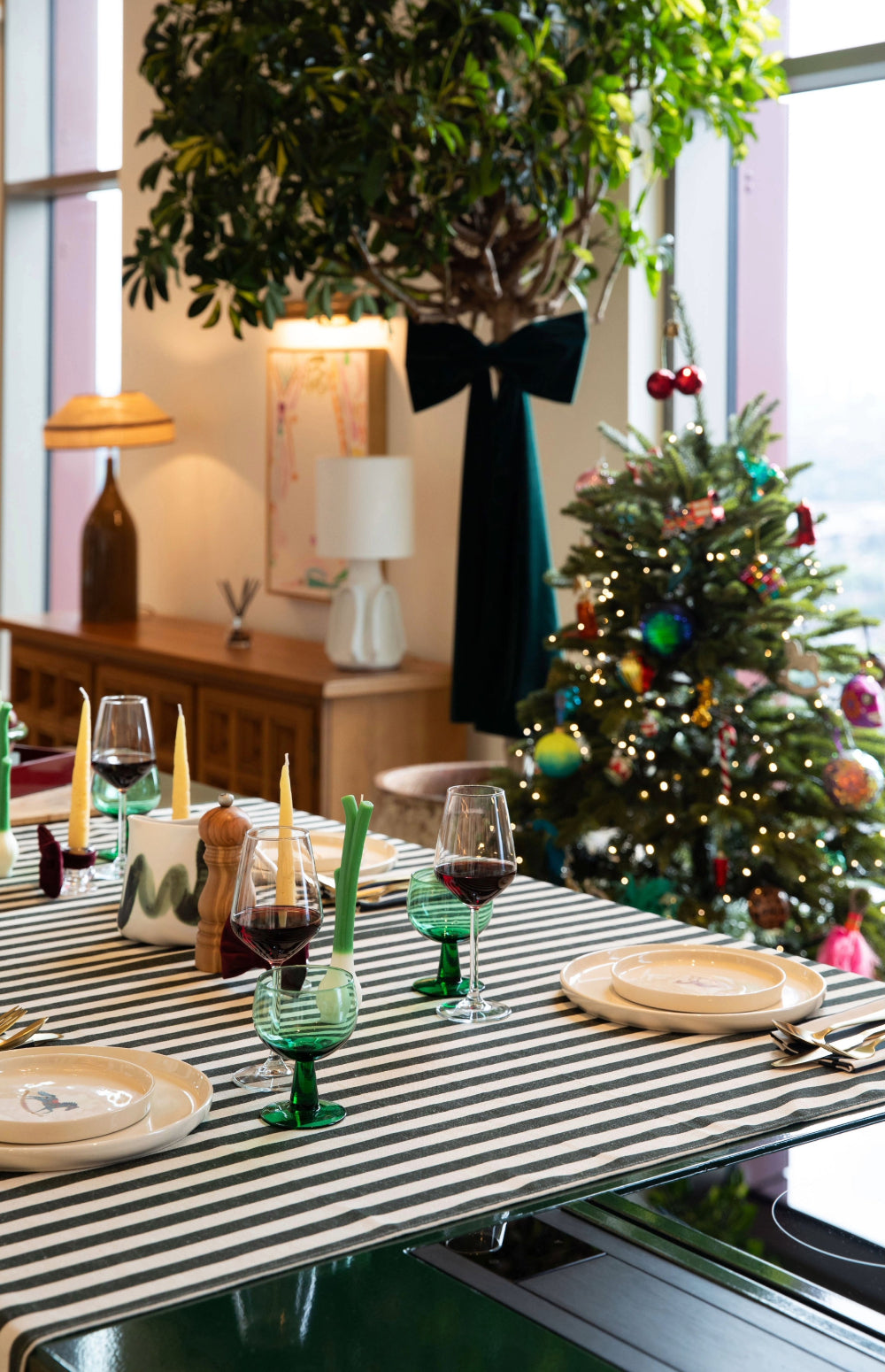 A festive holiday dining table setup in a decorated living space, featuring a black and white striped table runner, green stem candle holders, wine glasses, and a decorated Christmas tree in the background.