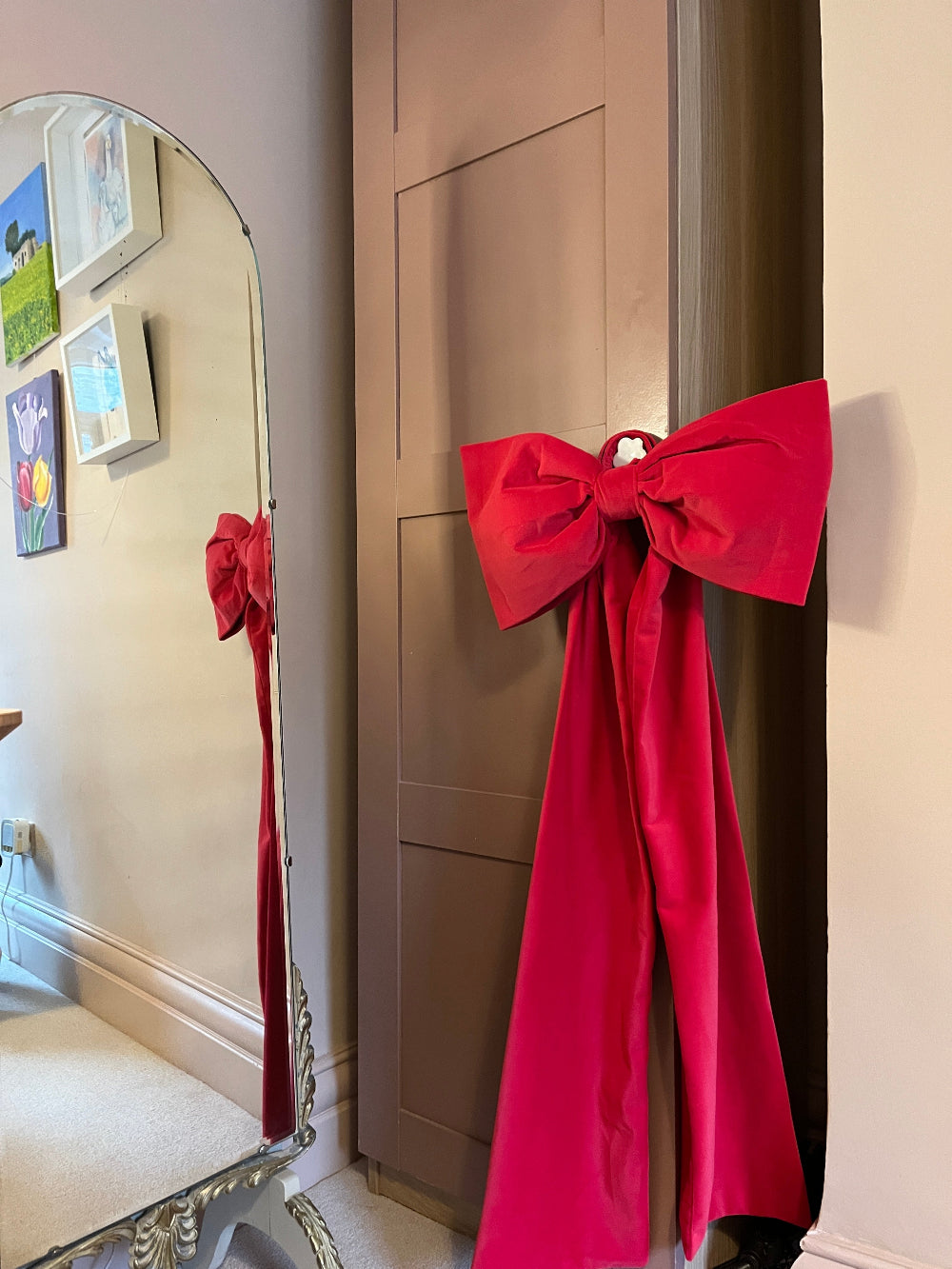 A bright red velvet bow tied around a brown wooden door in an indoor room, with a matching red ribbon hanging down the side of the door and a mirror on the left reflecting the bow.