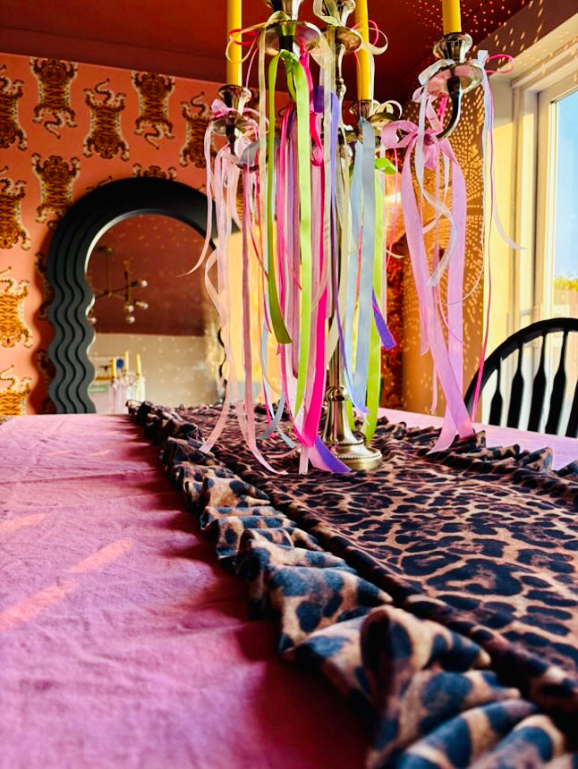A bohemian-style dining table centerpiece featuring a leopard-print runner draped over a pink tablecloth, with a multi-arm candelabra holding yellow candles and colorful hanging ribbons.