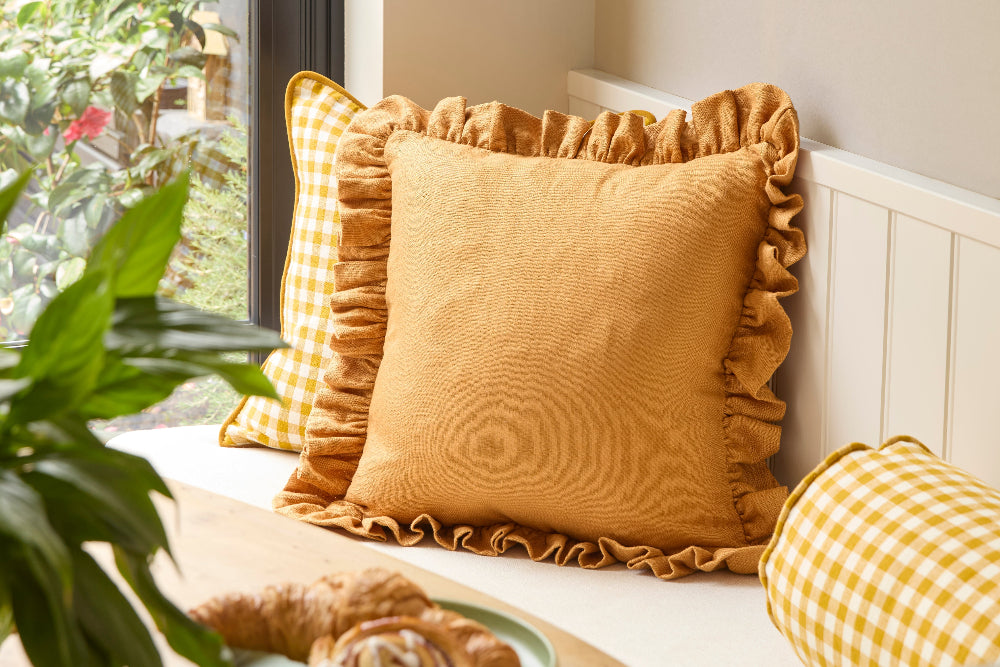 Orange cushion with ruffled edges on a white bench next to a window with yellow checkered cushions.