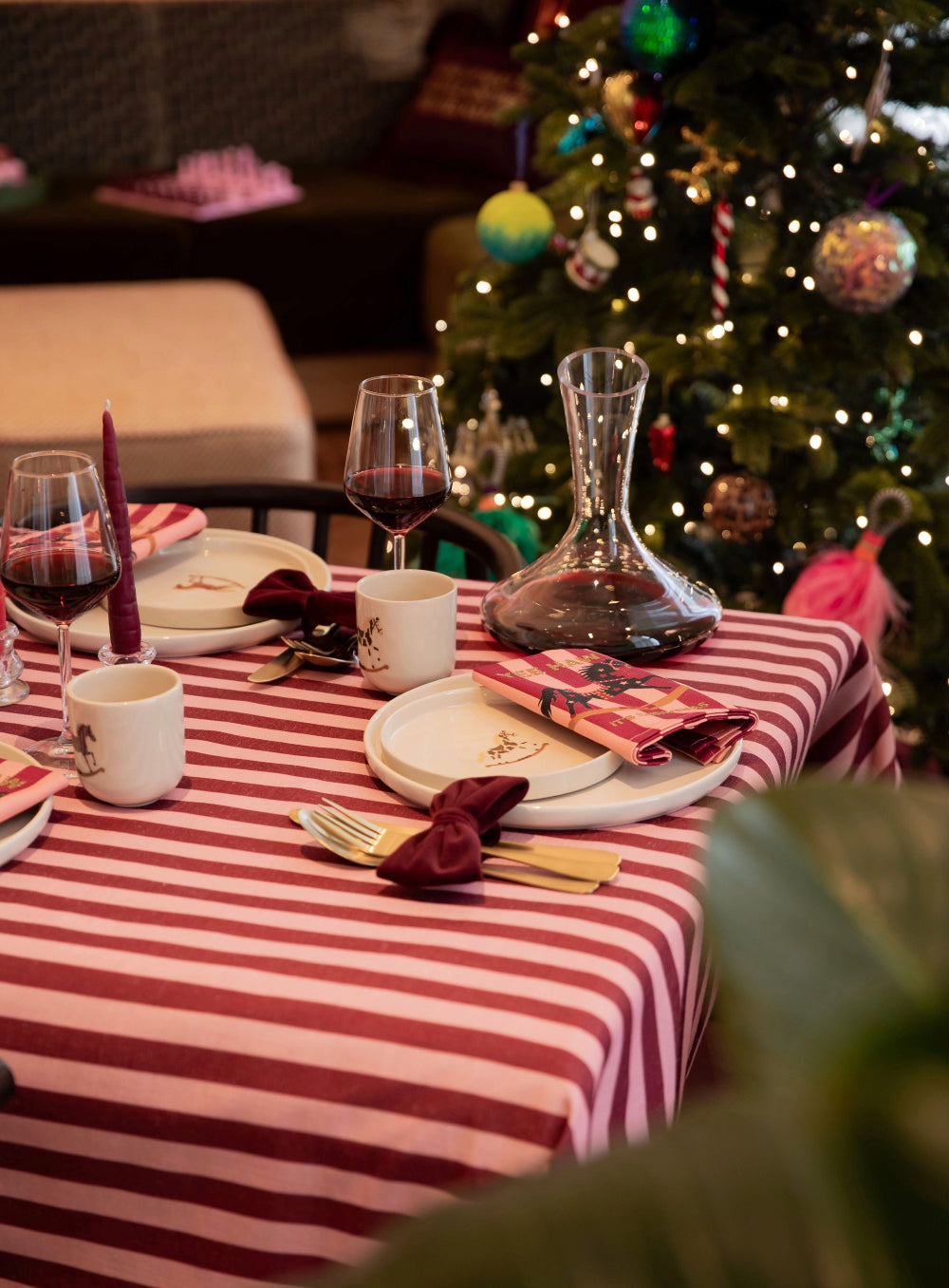 A festive holiday table setting with a red and white striped tablecloth, white plates, white mugs, wine glasses, gold cutlery, and burgundy napkins, set in front of a decorated Christmas tree.