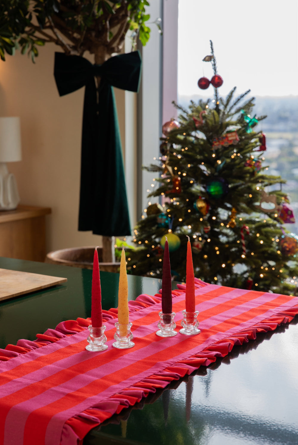 A festive holiday table setting featuring a red and pink plaid table runner with ruffled edges, decorated with four taper candles in red, yellow, and burgundy. In the background, a decorated Christmas tree stands near a window.