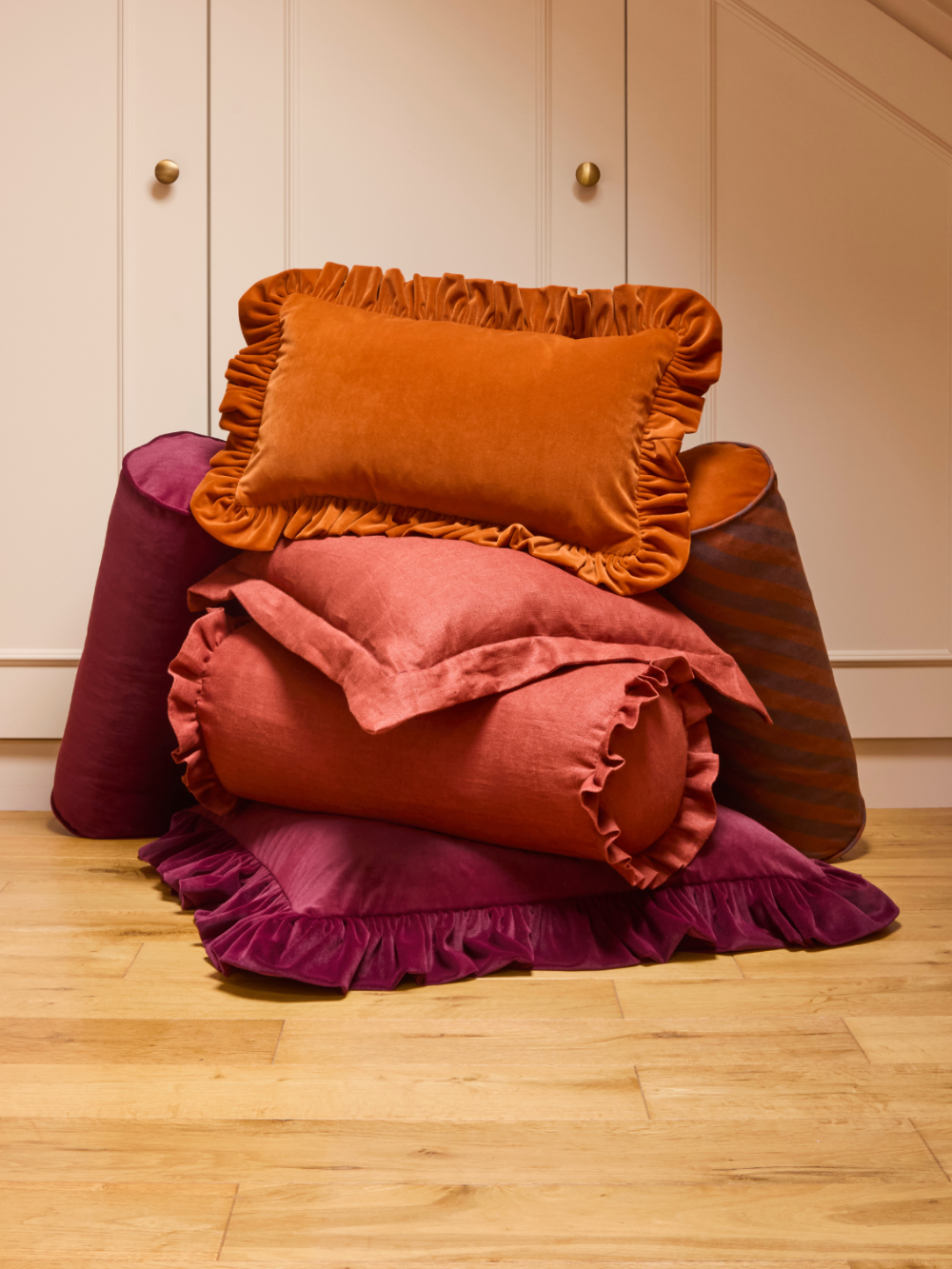 A styled collection of velvet ruffled pillows and cushions in warm autumn and deep berry tones, arranged on a light wood floor in front of cream-colored paneled walls.
