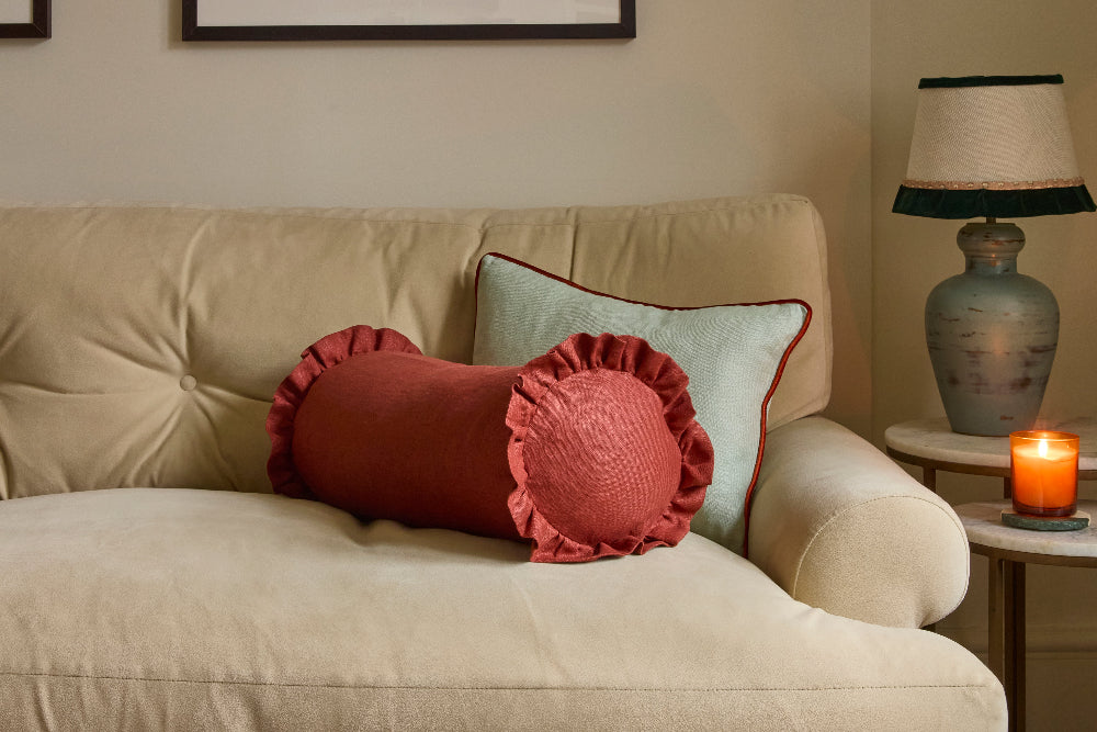 A styled living room scene featuring a beige sofa with two decorative pillows: a rust-red ruffled bolster pillow in the foreground and a sage green pillow with red piping in the background, next to a lit candle on a side table and a lamp.