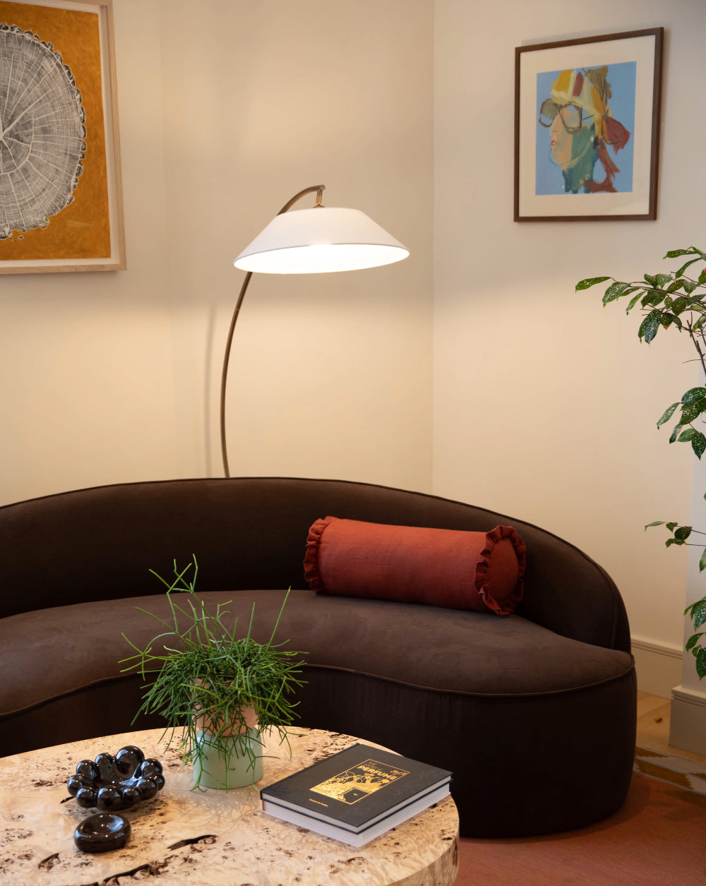 A curved dark brown upholstered sofa in a styled living room corner, with a rust-red cylindrical pillow, a white floor lamp, framed wall art, a plant, and a marble-top table in the foreground.