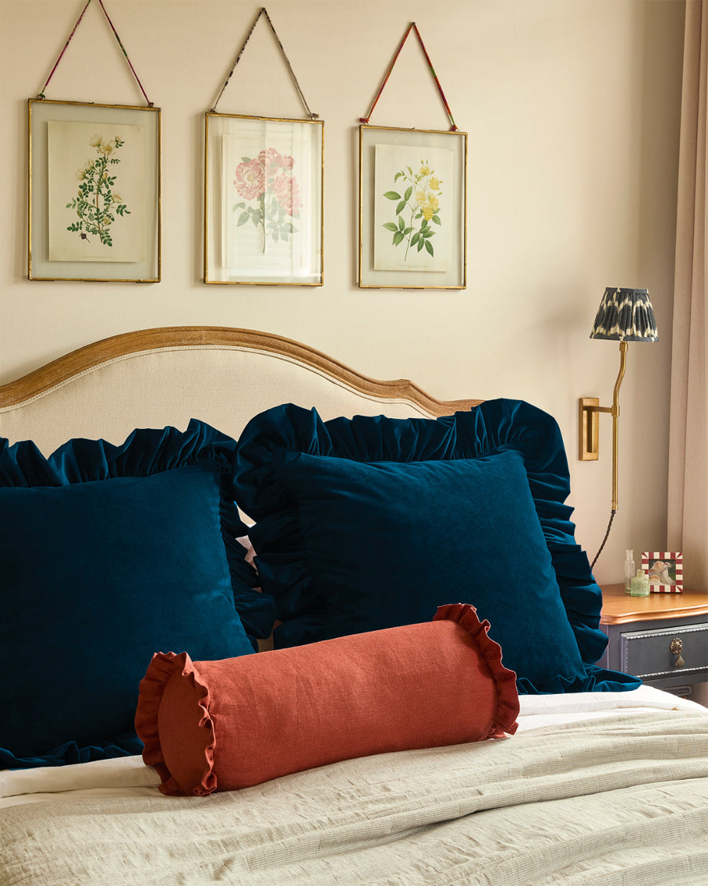 A styled bedroom scene featuring a bed with deep teal ruffled velvet pillows, a rust-orange ruffled cylindrical bolster pillow, and a cream-colored textured duvet. Above the bed, three framed botanical prints hang on a beige wall next to a gold wall lamp.