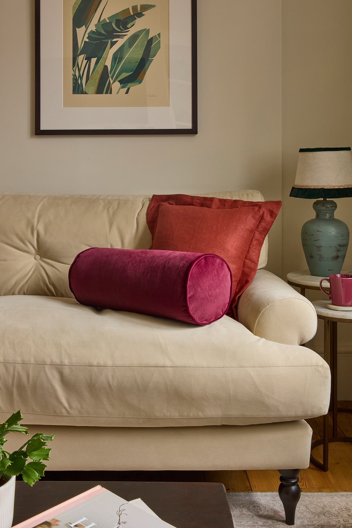 A styled living room scene featuring a cream-colored sofa with two decorative throw pillows: a cylindrical magenta velvet pillow in front and a rectangular rust-red pillow behind it. A framed botanical print hangs on the beige wall above, with a side table, blue-green lamp, and mug to the right.