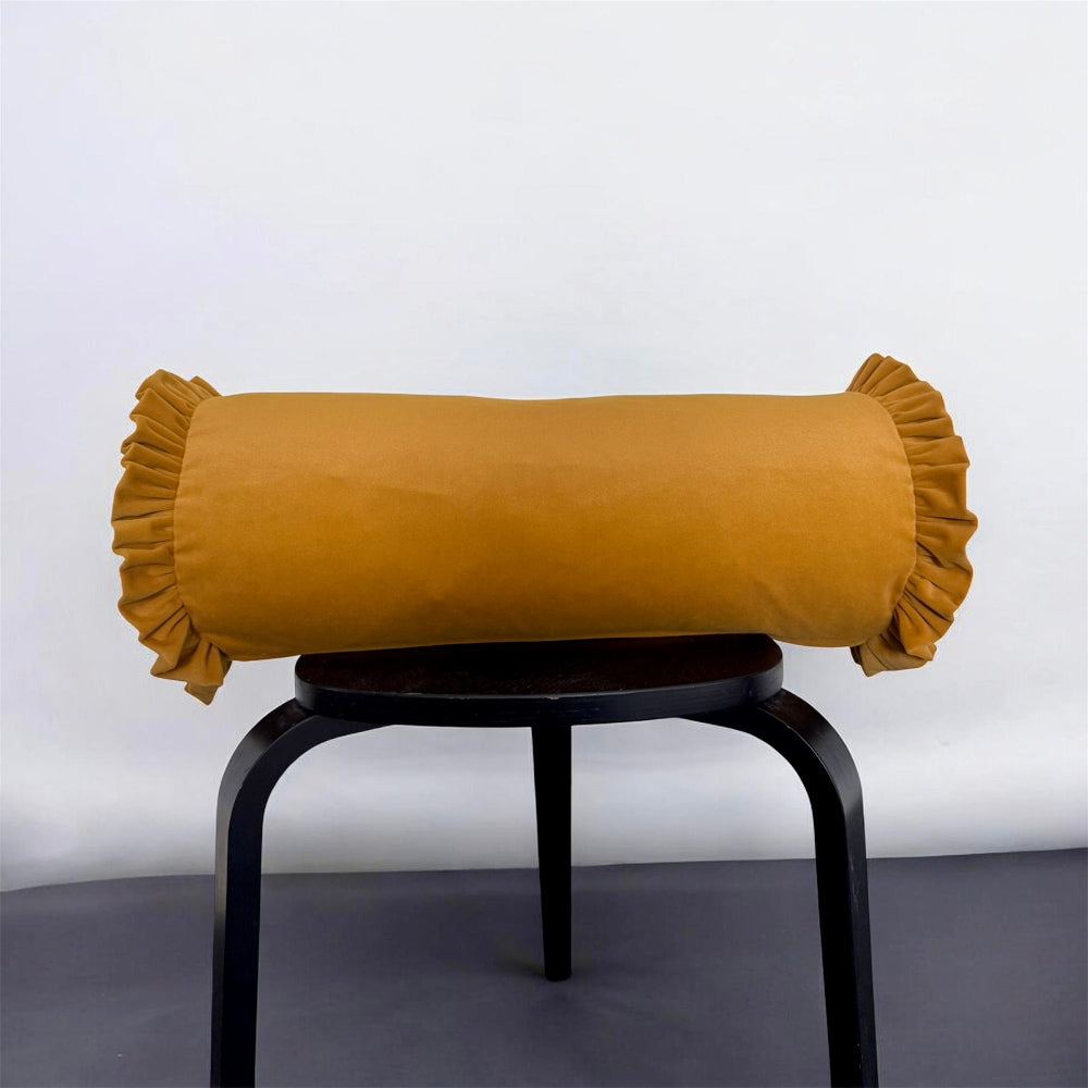 A mustard yellow cylindrical lumbar pillow with ruffled trim on both ends, displayed on a black metal stool against a white wall and dark floor.