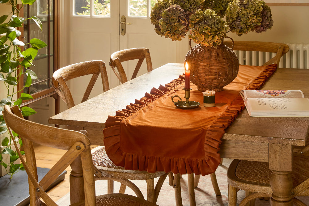 An orange ruffled table runner draped over a wooden dining table, styled with a wicker vase of dried flowers, a lit candle, and wooden chairs in a warm, sunlit room.