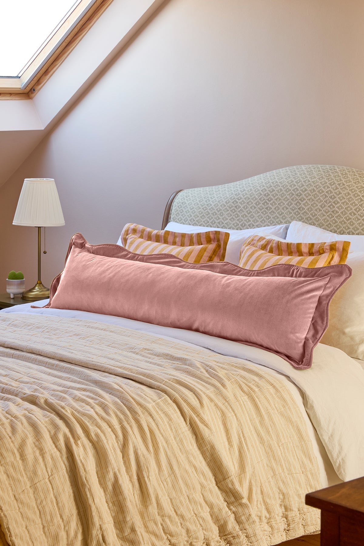 A bedroom scene showing a bed made up with a dusty pink lumbar pillow, striped yellow and white accent pillows, and a cream textured duvet. The background features a patterned headboard, a bedside table with a lamp, and a sloped ceiling with a skylight.