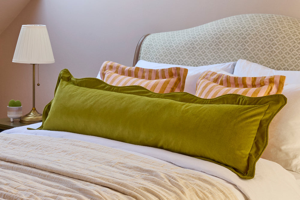 An olive green velvet lumbar pillow resting on a bed with white bedding, striped accent pillows, and a patterned headboard in a bedroom setting.
