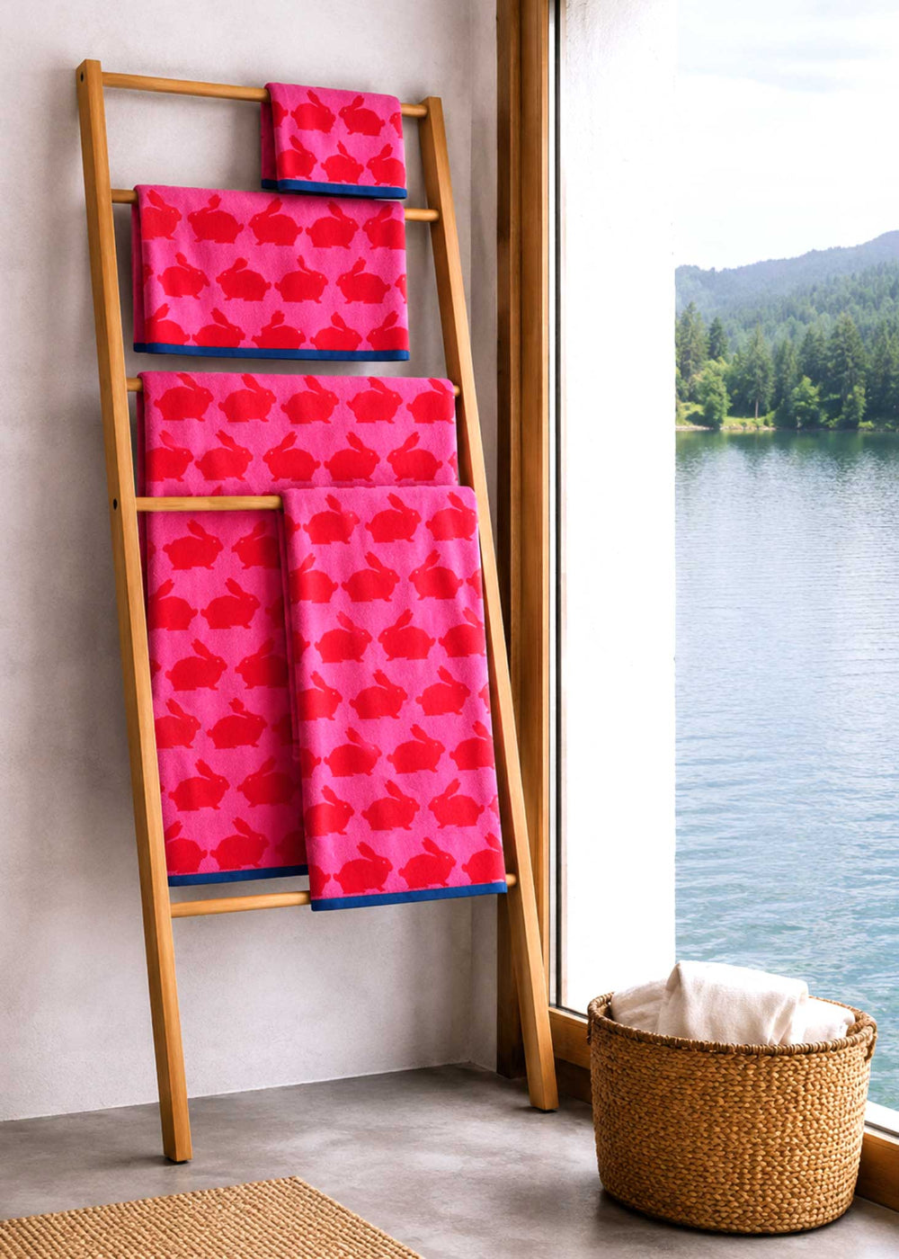 Pink patterned towels with rabbits on a wooden ladder by a window with a view of water and trees.