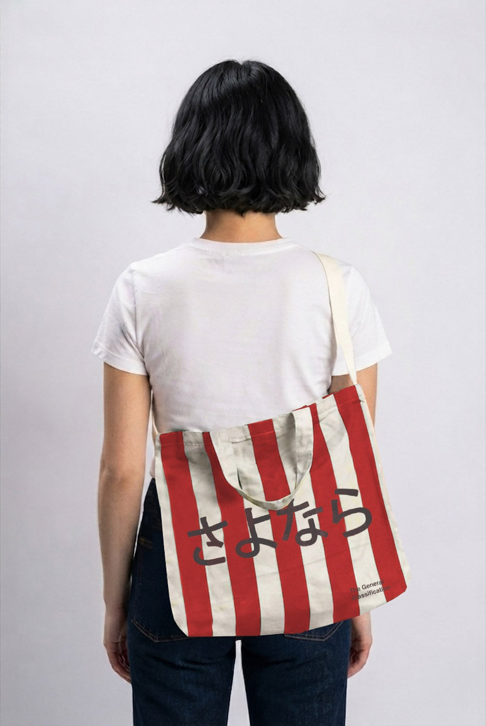 A person shown from behind wearing a white t-shirt and dark jeans, carrying a canvas tote bag over their shoulder. The bag features vertical red and off-white stripes with black Japanese text printed across the front.