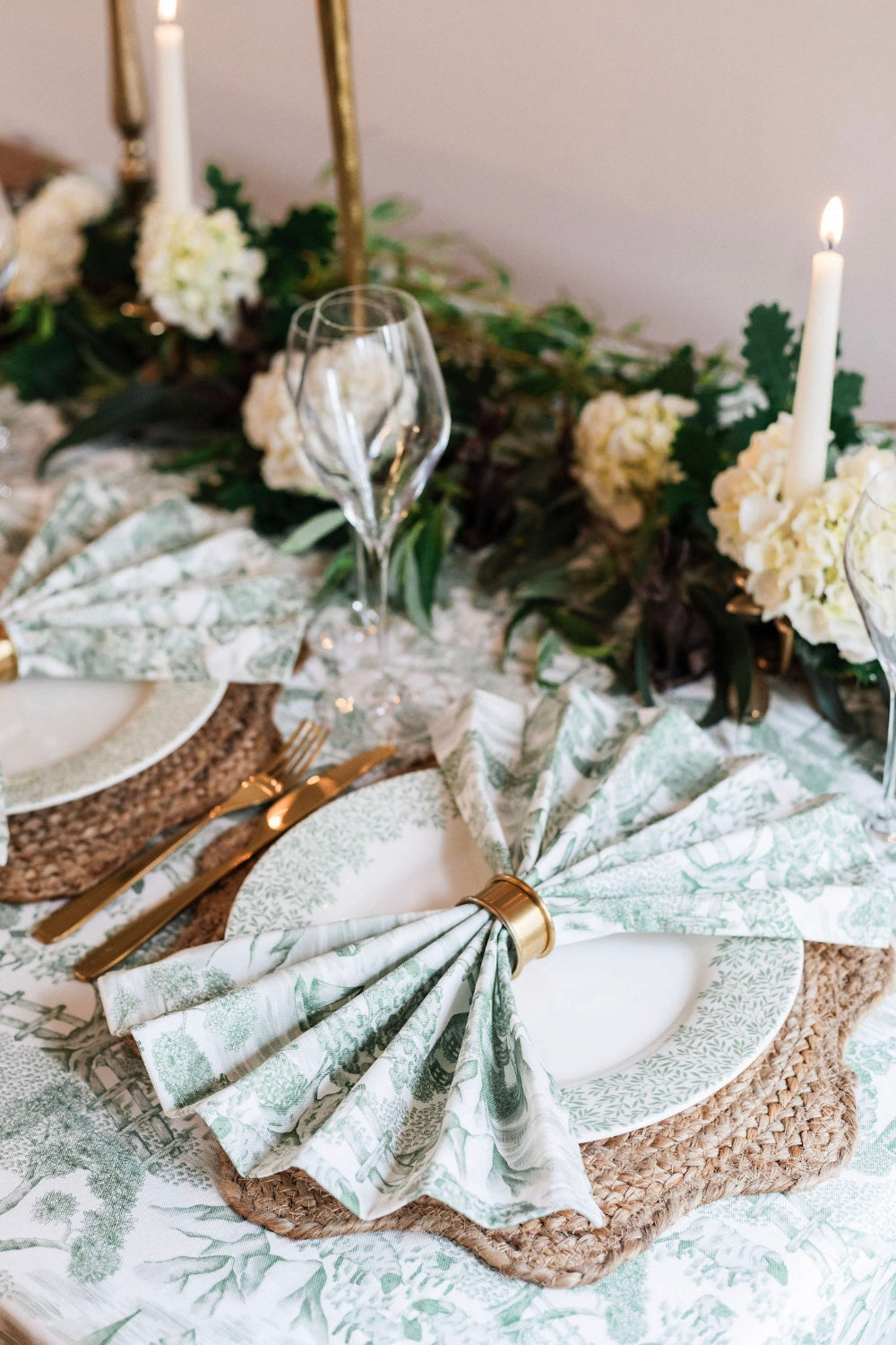 Elegant table setting with green and white floral napkins, gold cutlery, and white candles.