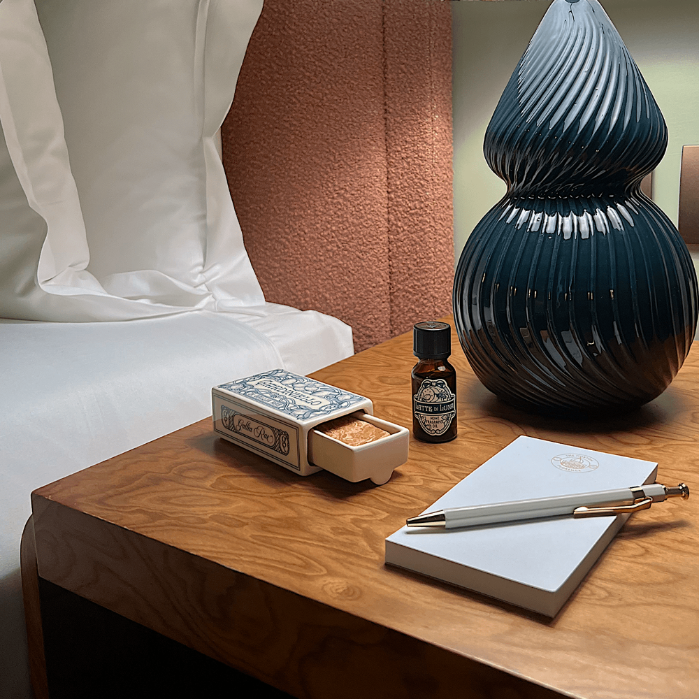 Wooden nightstand with decorative items including a vase, notebook, pen, and small ceramic matchbox with Gallia Rae home fragrance.