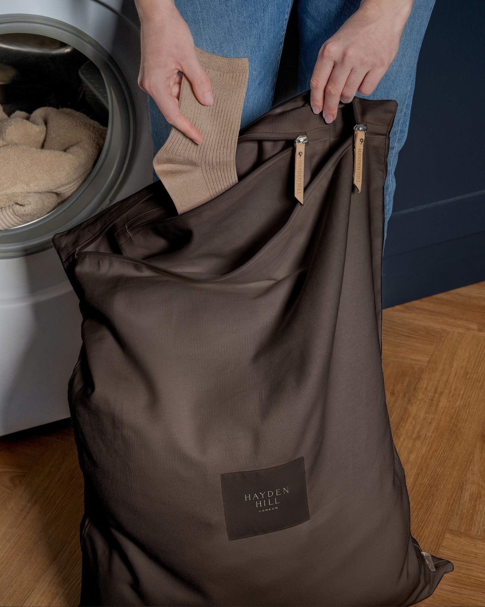 Person loading clothes into a brown laundry bag with 'Hayden Hill.' label next to a washing machine.
