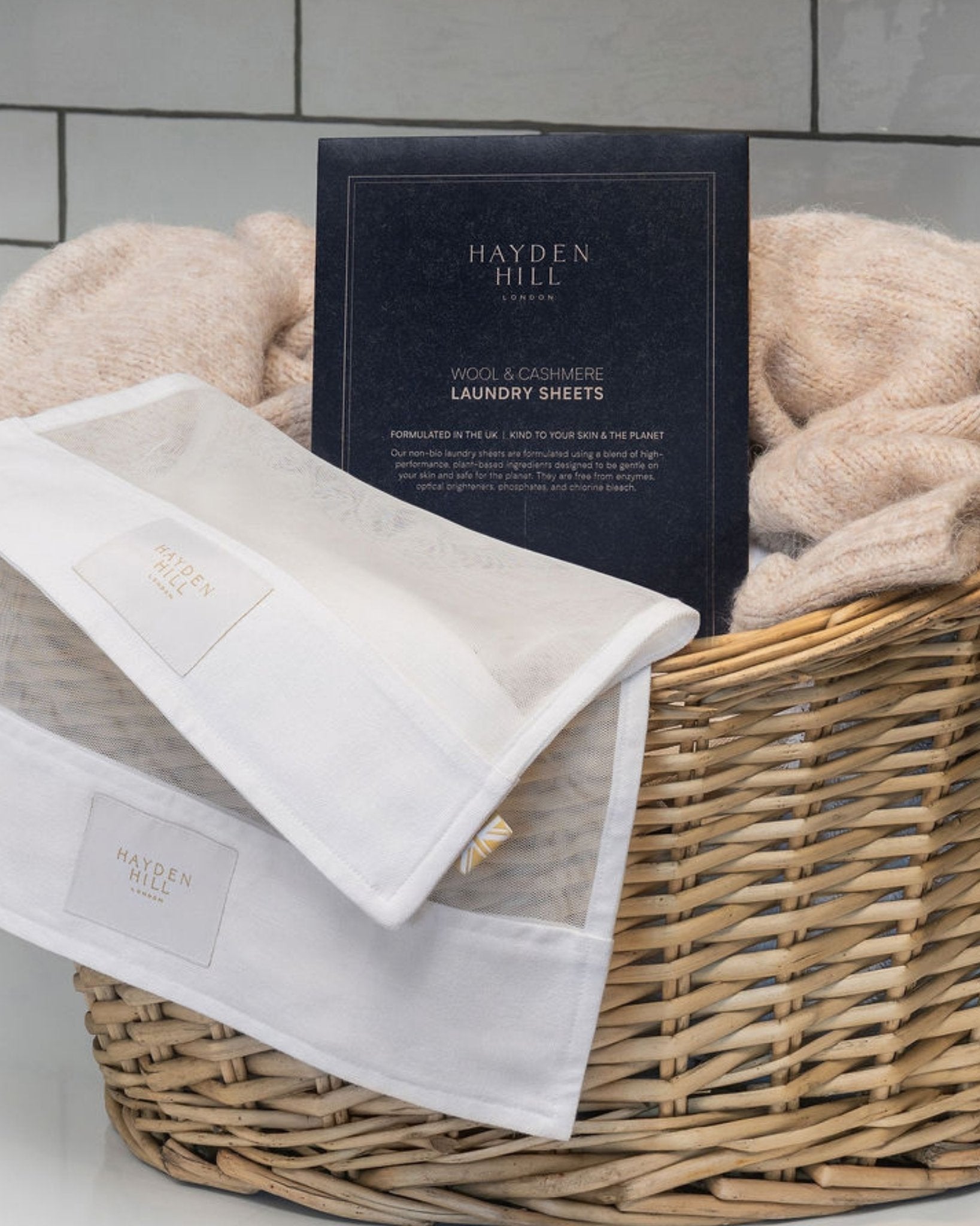 A wicker basket filled with soft beige towels and a navy Hayden Hill London box of wool-blend laundry sheets, set against a light gray tiled wall.