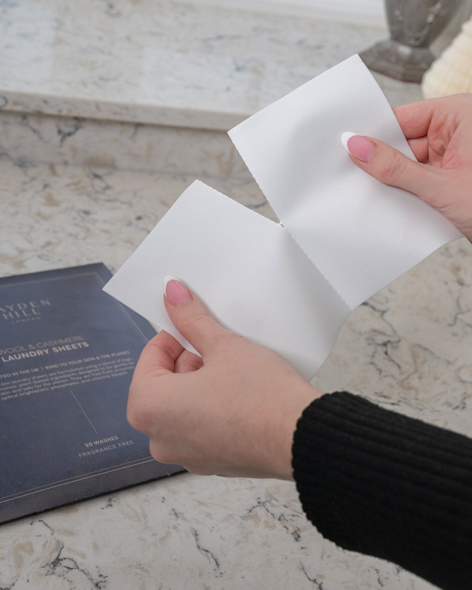 Hands holding white laundry sheets on a marble surface with a product box in the background.
