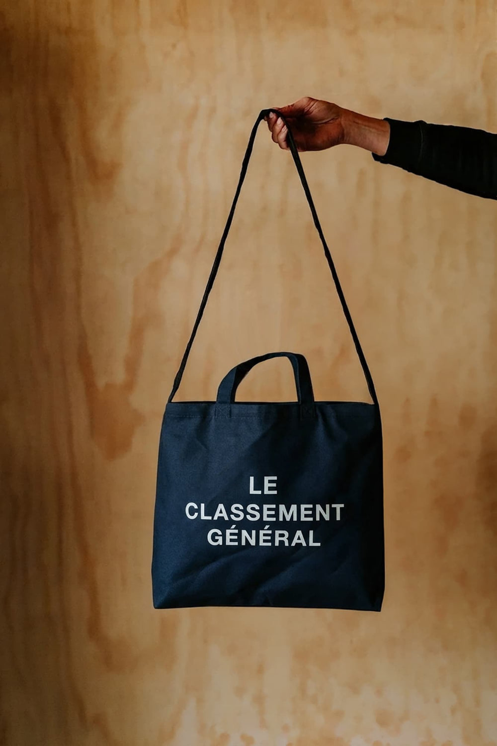 A navy blue canvas tote bag with white printed text reading 'LE CLASSEMENT GÉNÉRAL' is held by a person's hand against a warm wood-paneled background.