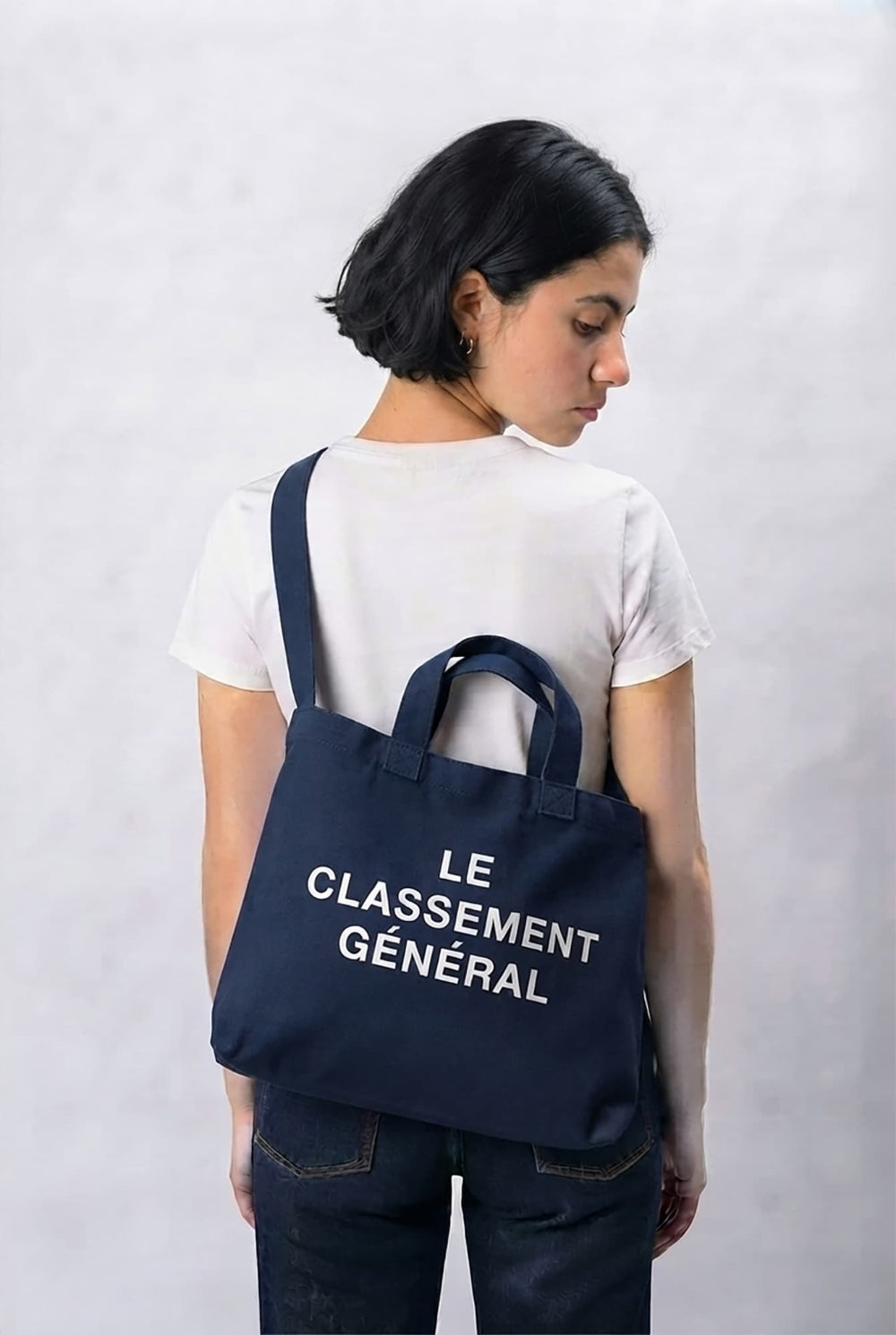 A woman wearing a white t-shirt and dark jeans is shown from behind, carrying a navy blue canvas tote bag over her shoulder. The bag features the white printed text 'LE CLASSEMENT GÉNÉRAL'.
