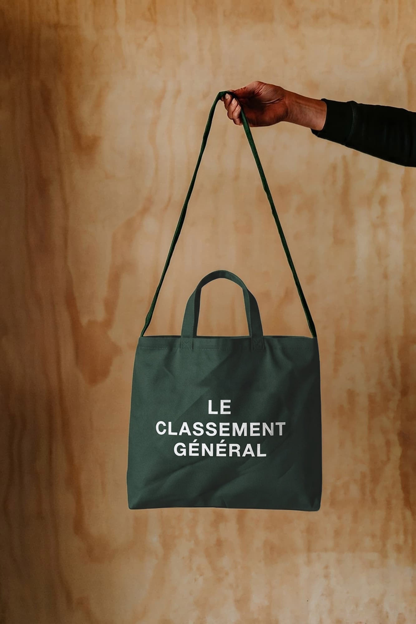 A dark green canvas tote bag with white printed text reading 'LE CLASSEMENT GÉNÉRAL' is held by a person's hand against a warm wood-panelled background.