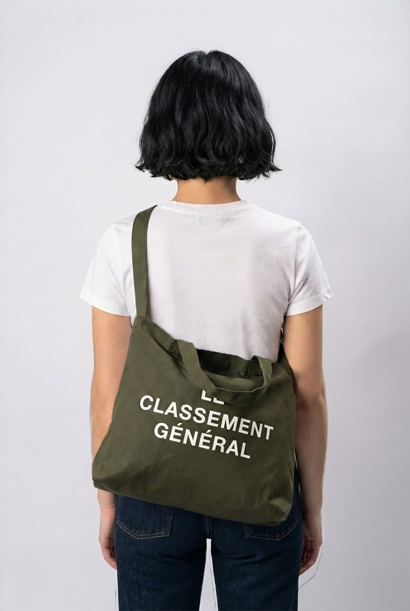 A person viewed from behind wearing a white t-shirt and dark blue jeans, carrying an olive green canvas tote bag over their shoulder. The bag features the white printed text 'LE CLASSEMENT GÉNÉRAL'.