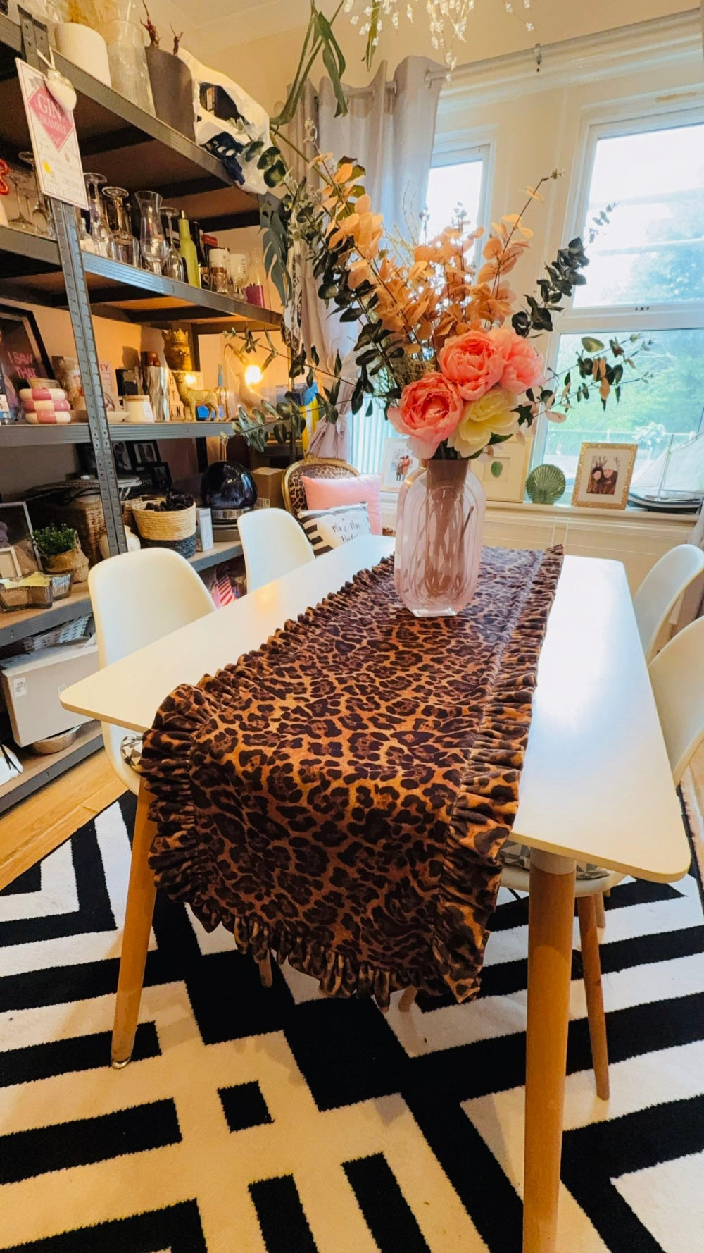 A leopard print throw blanket with fringe is draped over a white dining table in a bright, decorated room. A pink vase filled with orange and pink flowers sits on top of the table, surrounded by white chairs.