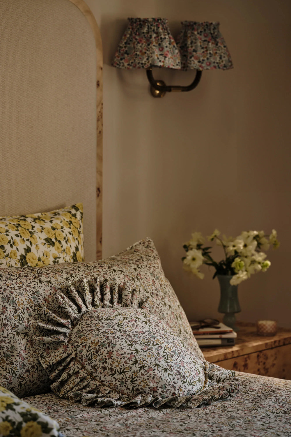 Floral-patterned pillows on a bed with a wall lamp and vase in the background.