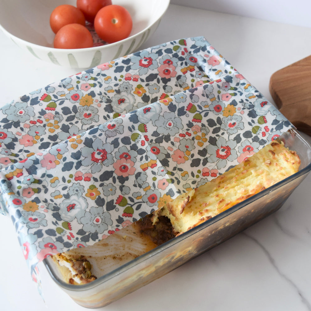 Glass dish with lasagna covered by a floral patterned liberty beeswax wrap, bowl of tomatoes in the background.