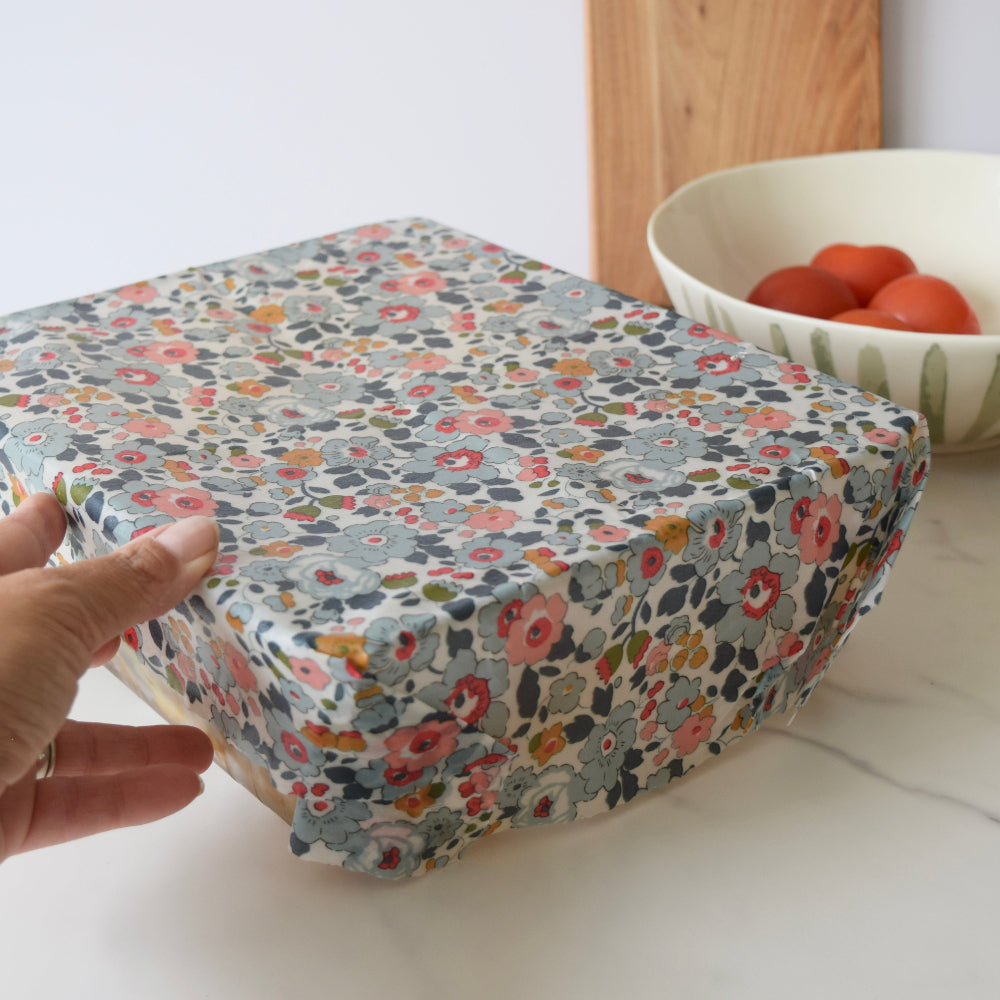Rectangular oven dish covered with a floral patterned beeswax wrap, held by a hand on a white surface.