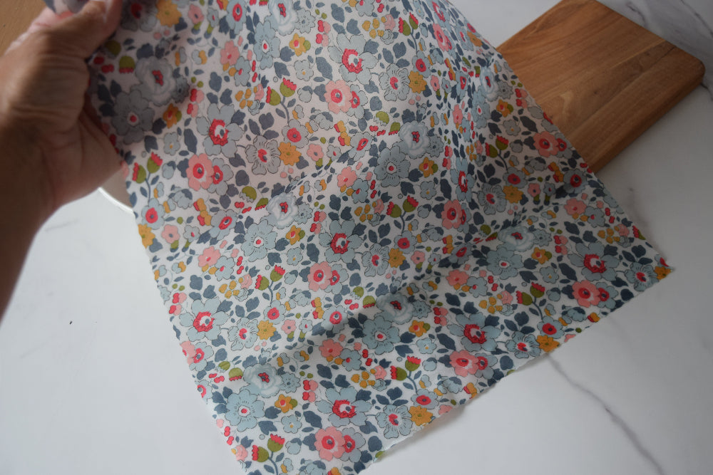Hand holding a piece of floral fabric beeswax wrap on a white surface with a wooden board.