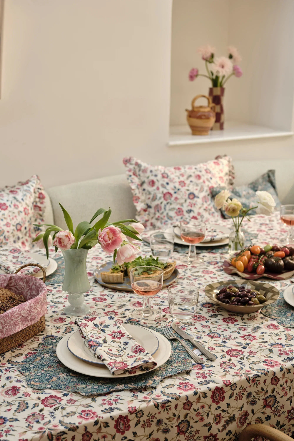 Dining table set with floral tablecloth, plates, glasses, and flowers in a cozy room.
