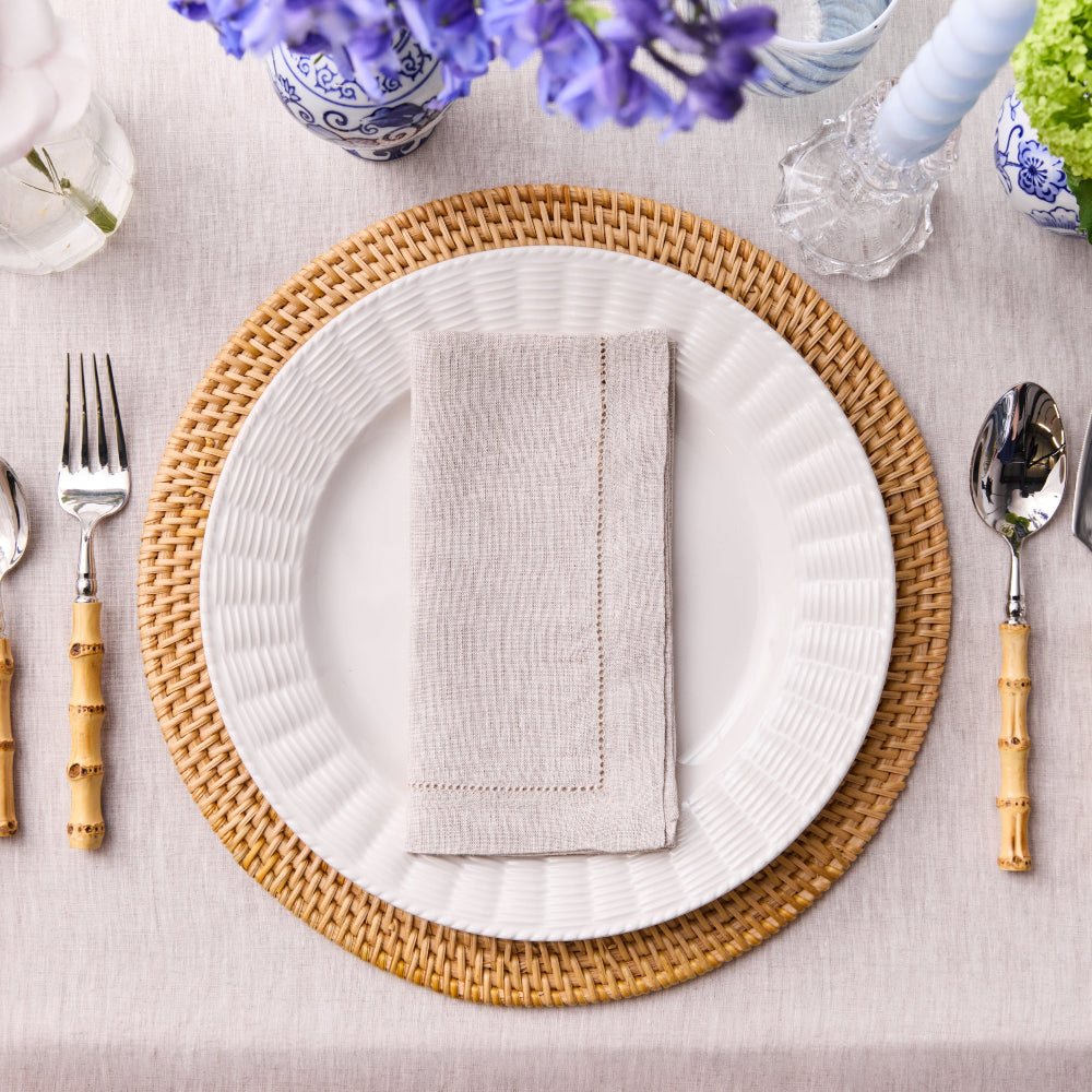 Dining table setting with hemstitched natural coloured napkins, white plates, woven placemats, and silverware on a light surface.