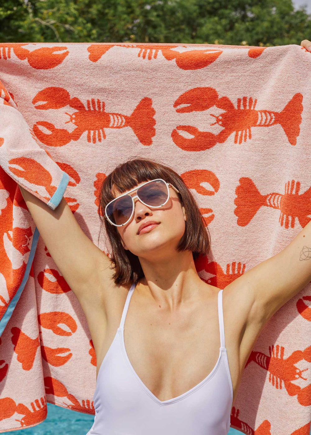 Woman in a white swimsuit and sunglasses lying on a towel with red lobster pattern.