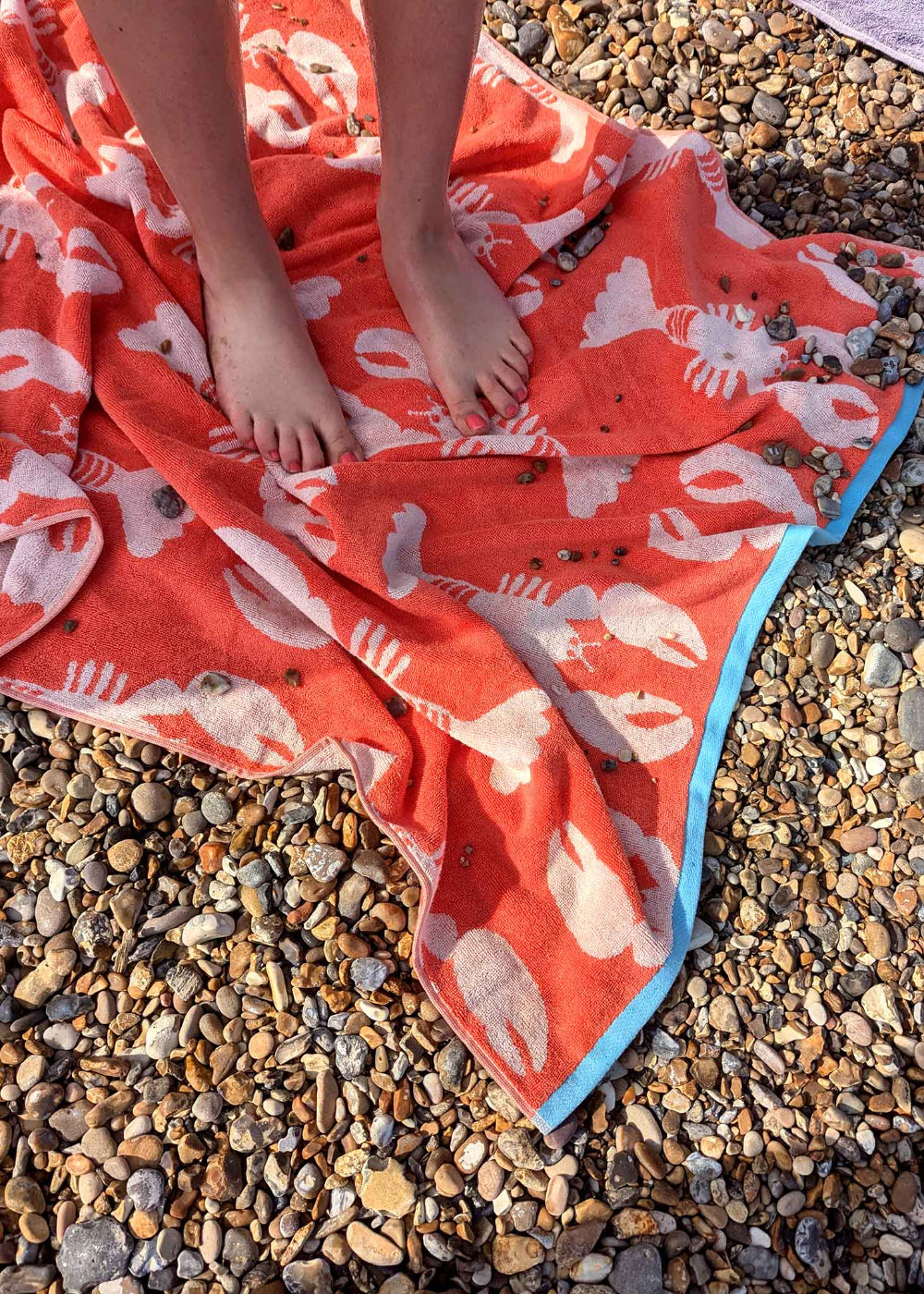 Person sitting on a red towel with white lobster pattern on pebbly ground