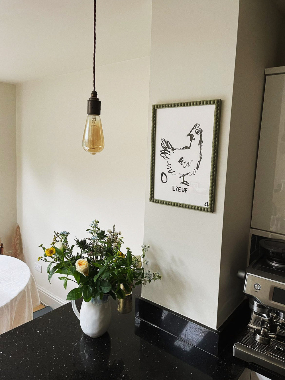 Modern kitchen interior with a vase of flowers on the counter, framed artwork on the wall, and a hanging light bulb.