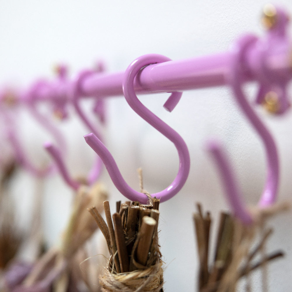 A close-up of a lavender purple metal rod with hanging S-hooks, with dried reeds or straw bundles suspended from some hooks against a plain light background.