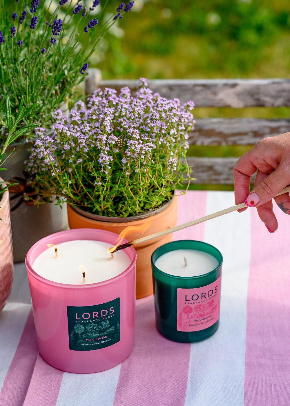 Two candles from Lords of the Wax being lit on a striped tablecloth with plants in the background.