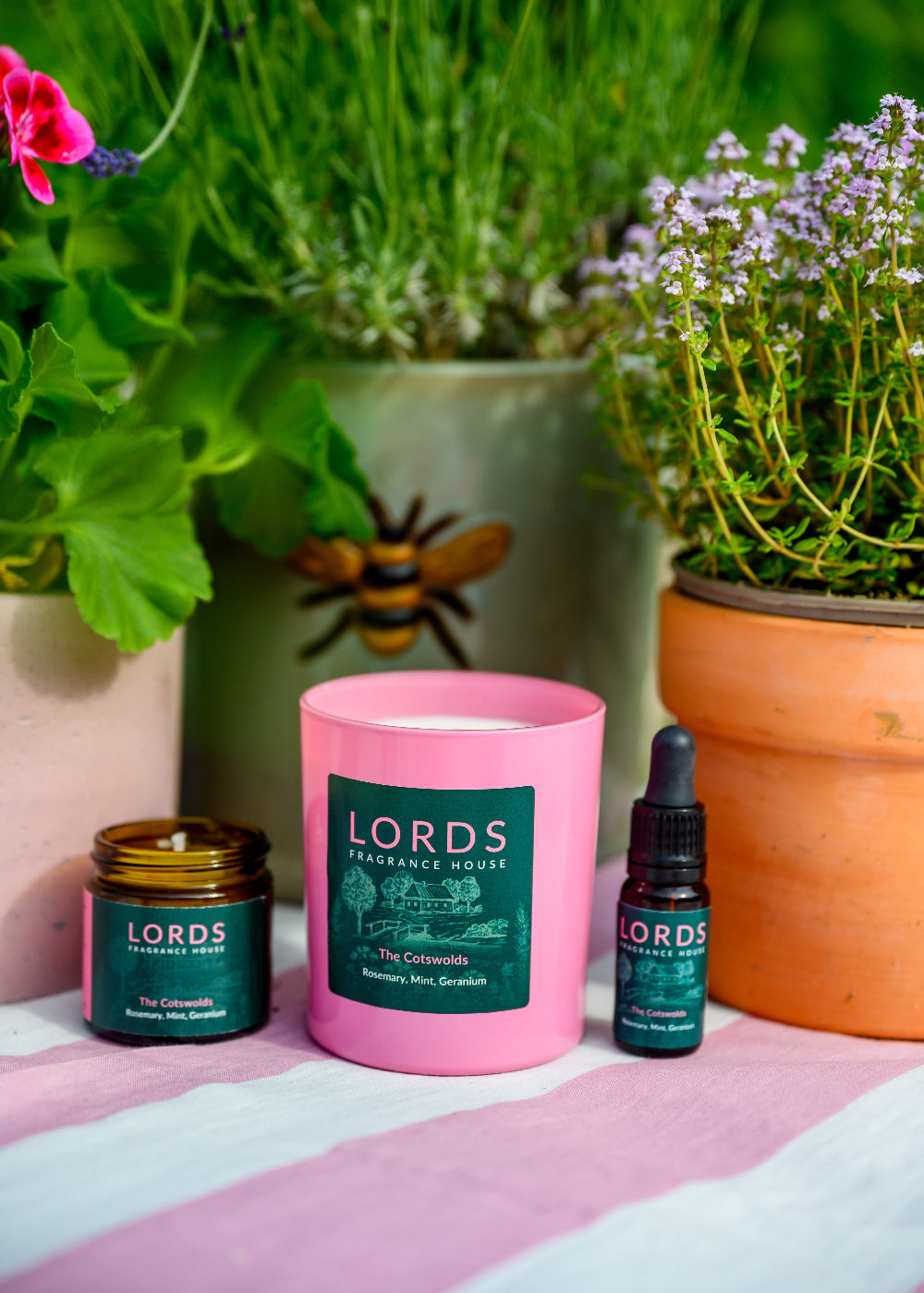 Lords cotswold products including a pink candle, green bottle, and black dropper bottle with plants in the background.