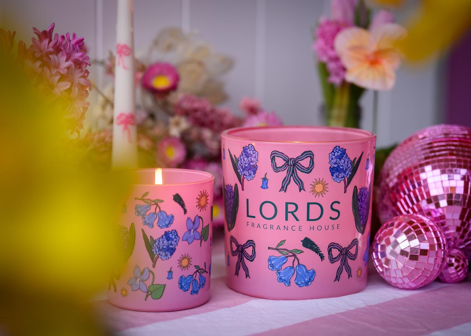 Two pink candles with floral designs on a surface, surrounded by flowers and disco balls.