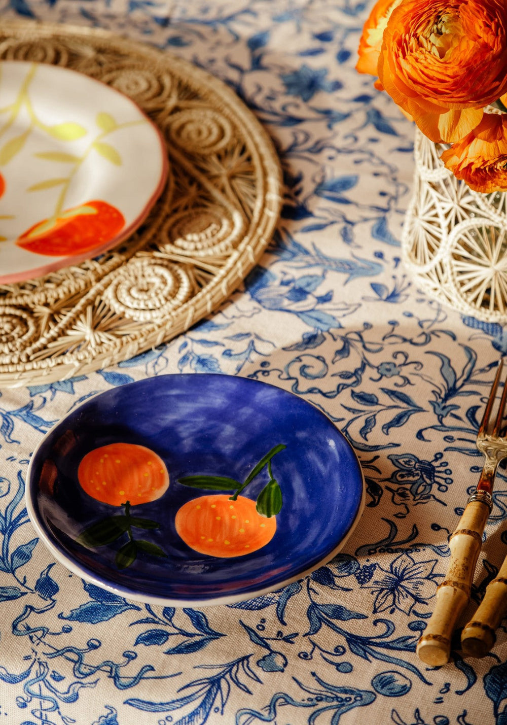 Decorative table setting with a blue plate featuring orange illustrations, a white plate with orange design, and a vase with orange flowers on a patterned tablecloth.