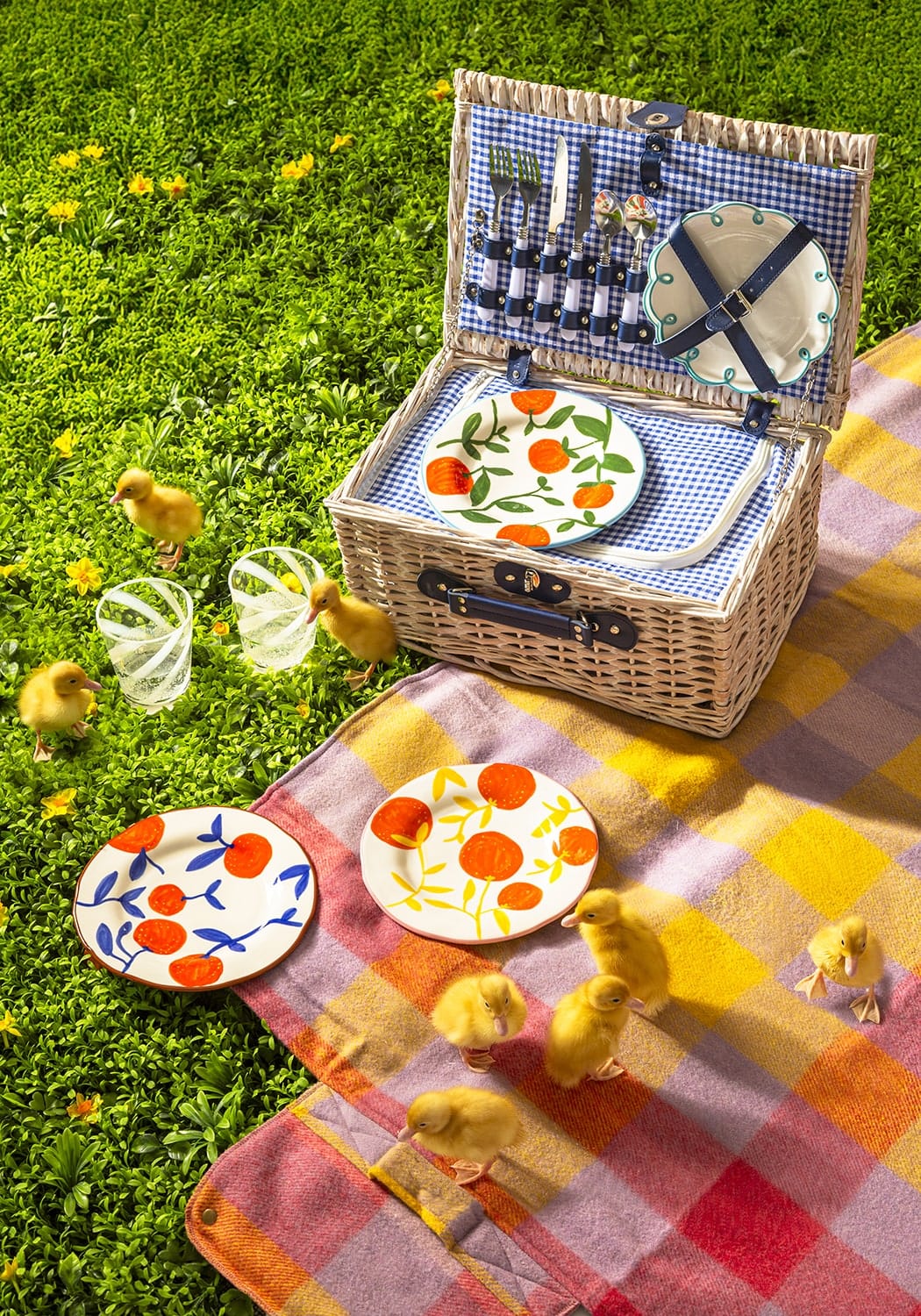 Open picnic basket with plates and glasses on a colorful blanket with chicks around, on grass.